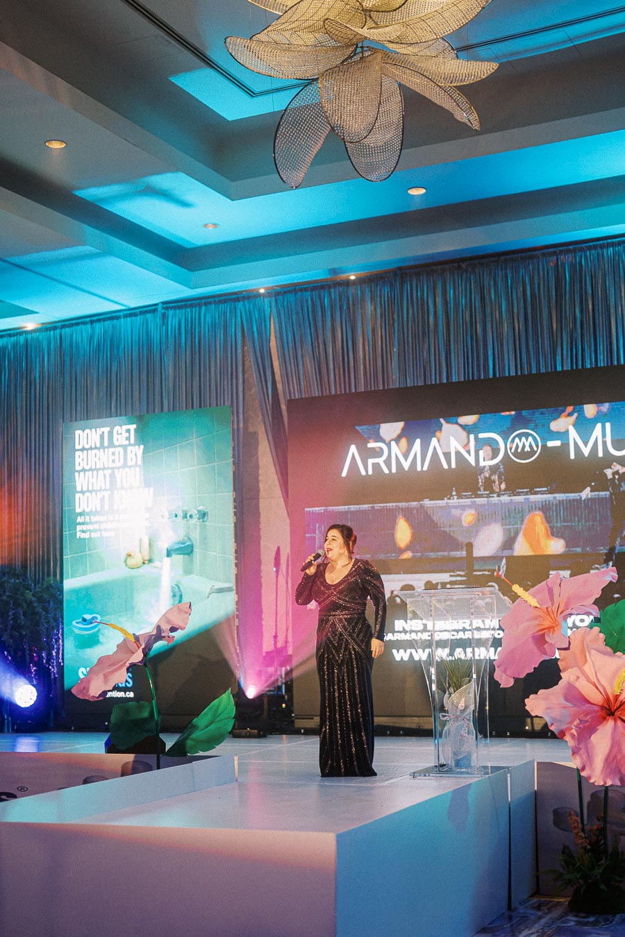 A woman in a glittering evening gown speaks into a microphone on stage, surrounded by floral decorations. A vibrant backdrop displays the name Armando with social media and website information. The event features colorful lighting and a modern ceiling design.