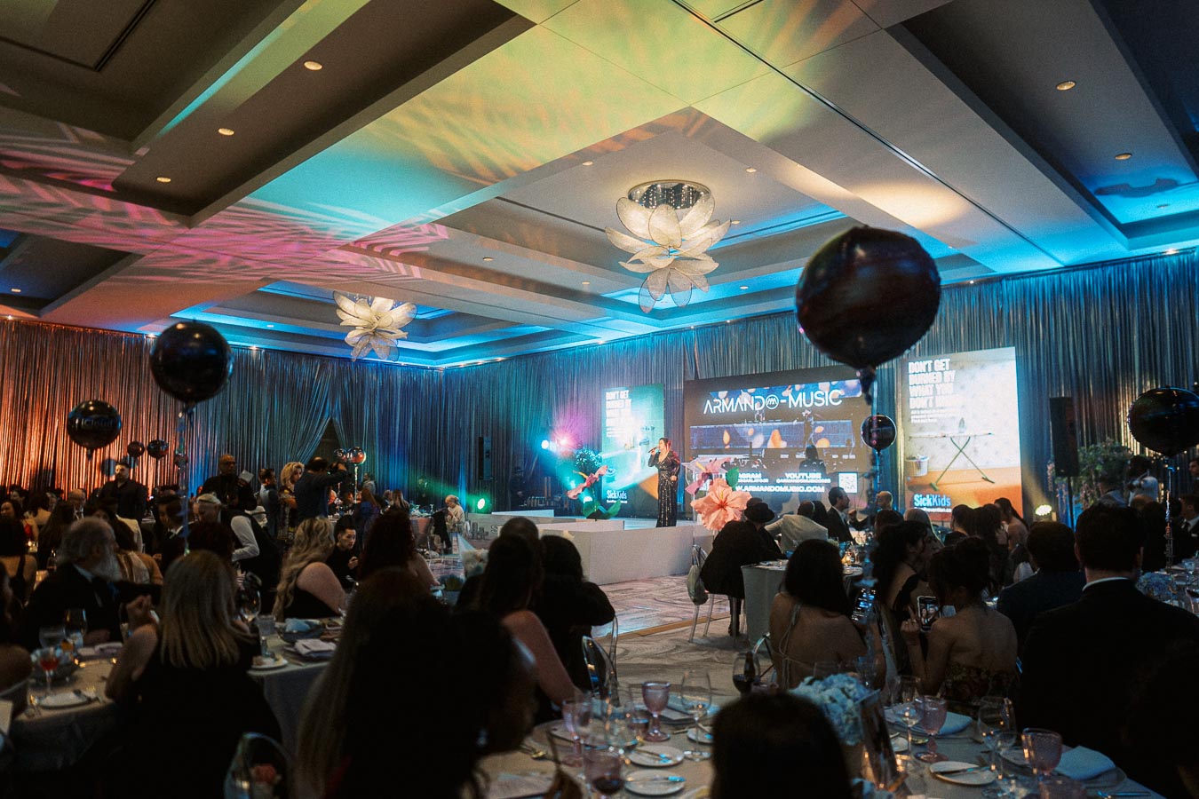 A lively gala event with guests seated at elegantly decorated tables, enjoying a musical performance on stage with colorful lighting and a vibrant atmosphere.