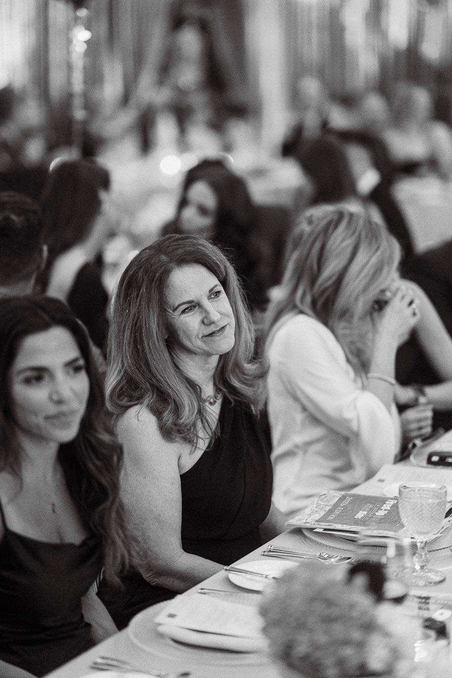 A black and white photo of an elegant woman in a one-shoulder dress attending a formal event. She sits at a beautifully set table, surrounded by other guests. The setting conveys a sophisticated and upscale atmosphere.