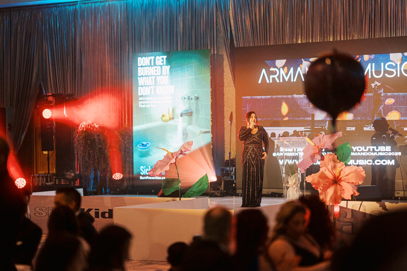Woman in an elegant black dress speaks at a glamorous event on stage with floral decorations and colorful lighting. A screen displays promotional content in the background.
