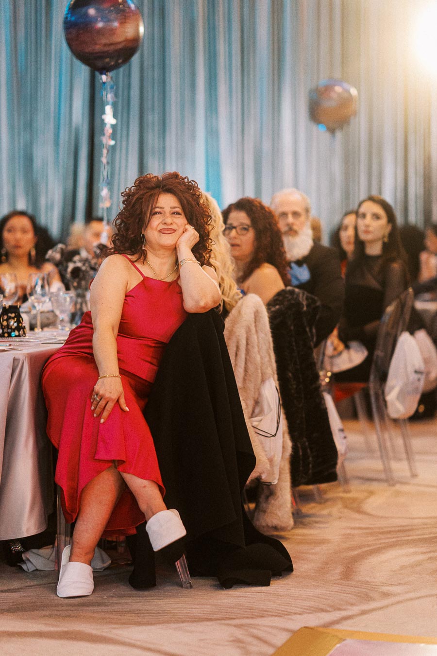 A woman in a red dress sitting at a formal event, smiling confidently with a balloon in the background, surrounded by other attendees.