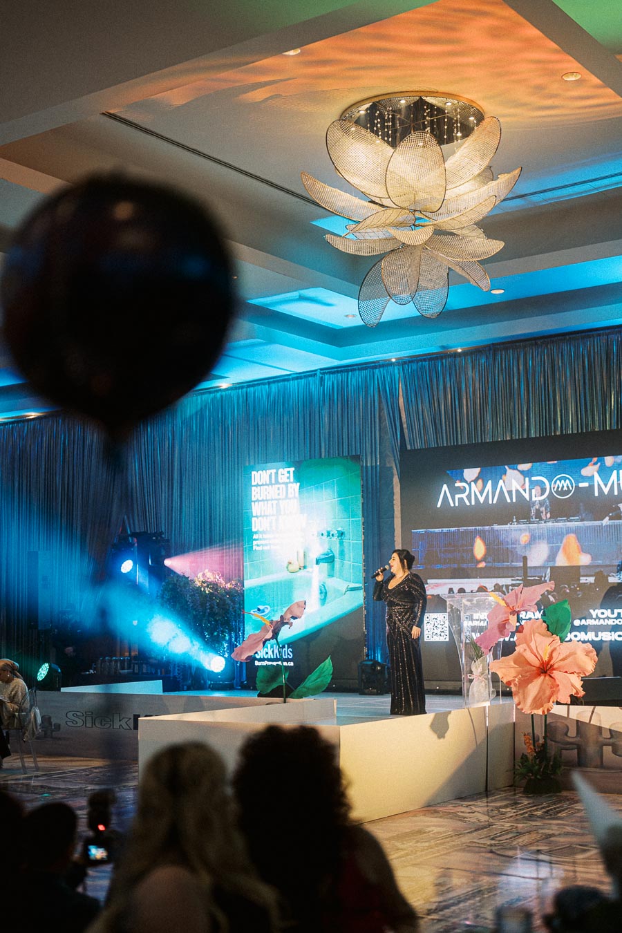 Singer performing on stage at a vibrant event with colorful lighting and large floral decorations, under an elegant ceiling chandelier.