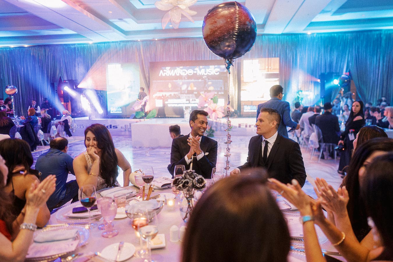 Guests seated at a glamorous event, with elegant decor, colorful lighting, and a large balloon centerpiece. People are dressed in formal attire, engaged in conversation and clapping, while a stage with bright screens is visible in the background.
