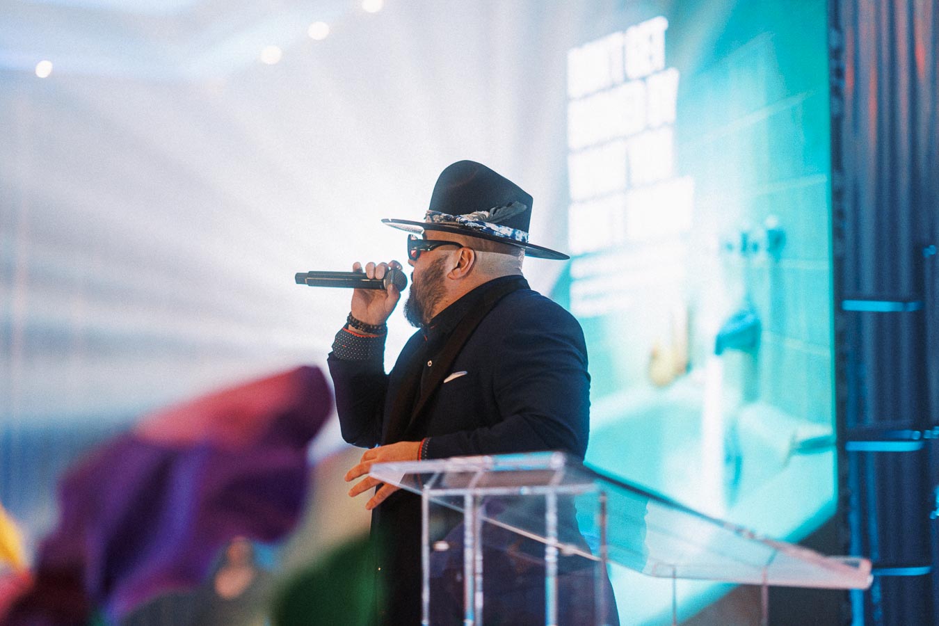 Man in stylish hat and sunglasses singing into microphone on stage with colorful background.