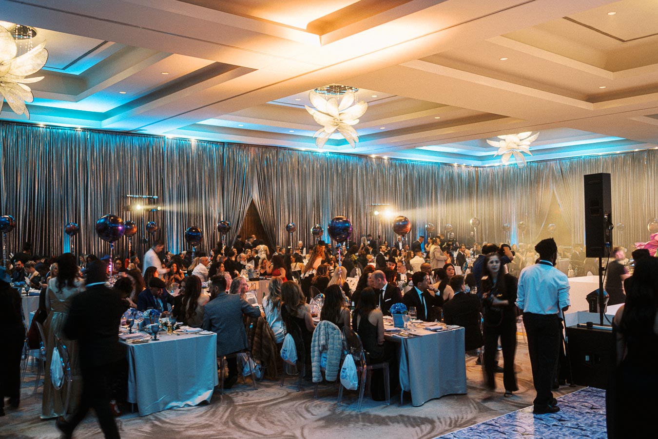 A vibrant and elegantly decorated banquet hall filled with well-dressed guests seated at tables, illuminated with atmospheric lighting and adorned with modern decorations and floral arrangements.