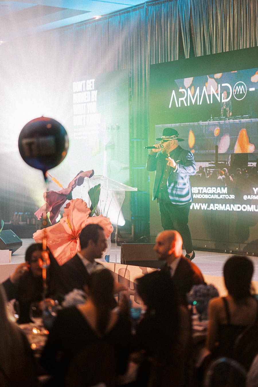 Live music performance at an elegant event with guests seated at tables and a male performer wearing a hat and suit on stage. The background displays a multimedia presentation with vibrant lighting.