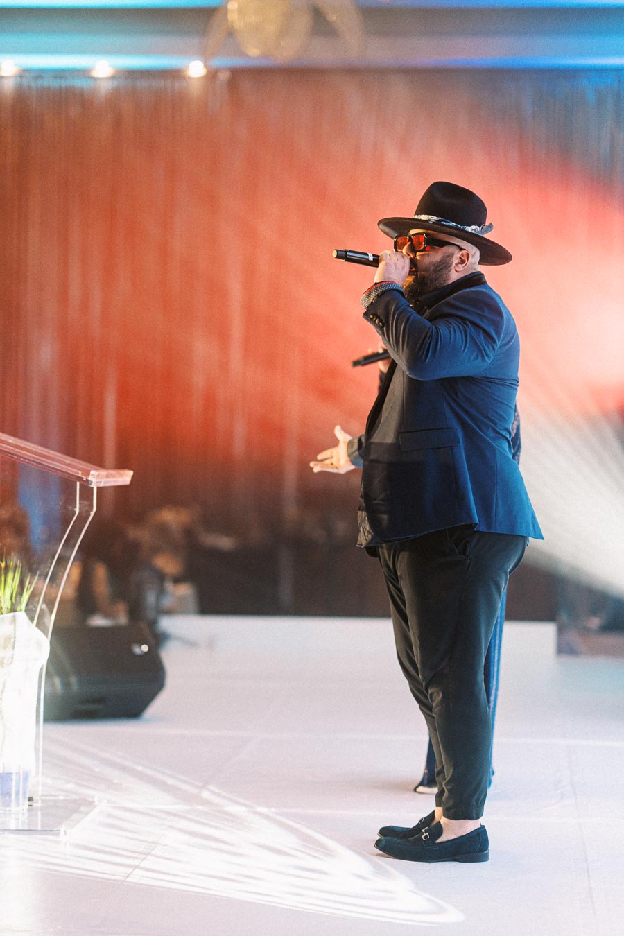 Man in stylish outfit and hat performing on stage with a microphone, vibrant lighting and audience in the background.