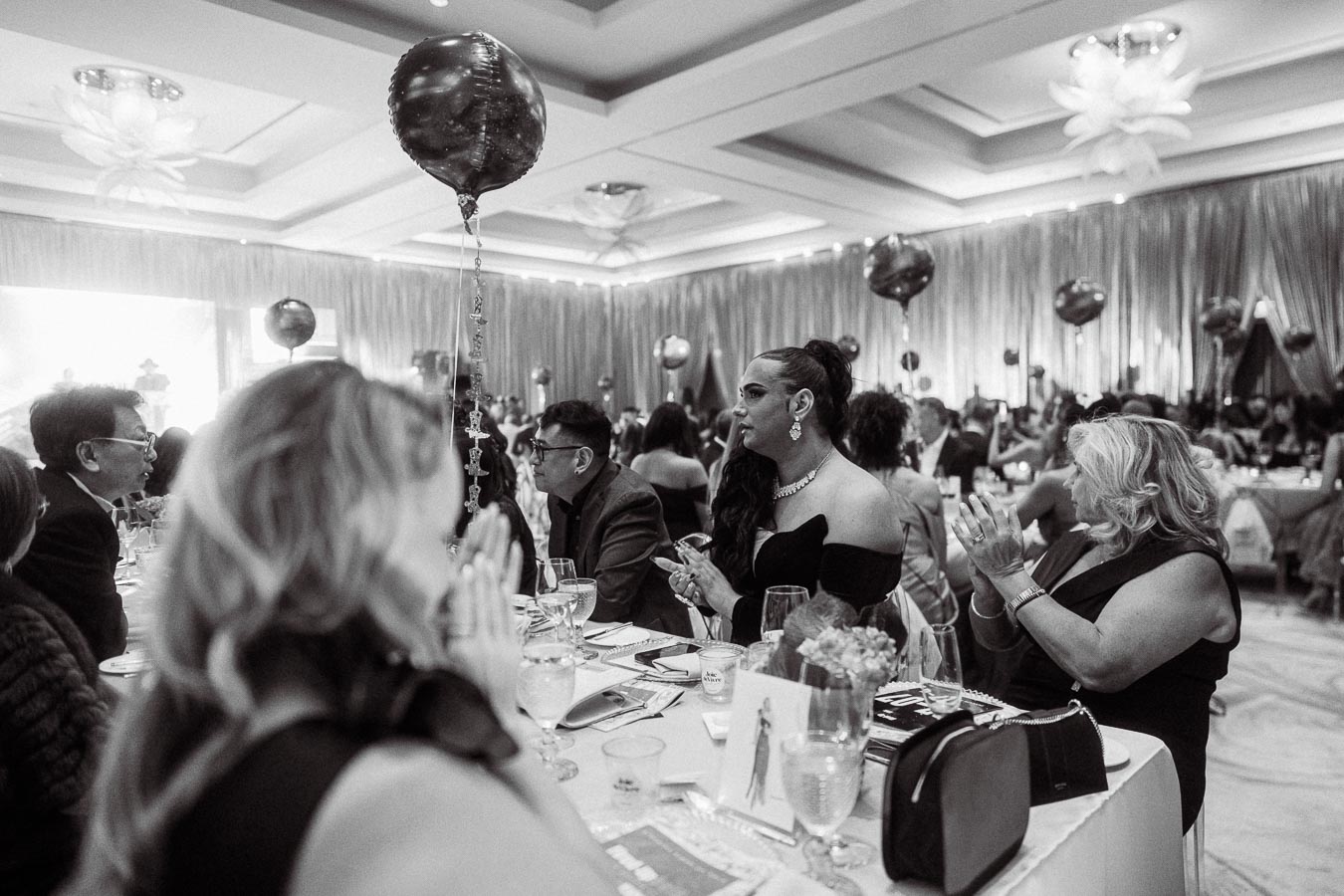 Black and white image of an elegant event, featuring guests dressed in formal attire seated at decorated tables, with attention drawn to the woman in the foreground clapping, under a ceiling adorned with balloons and shimmering drapes in a spacious venue.