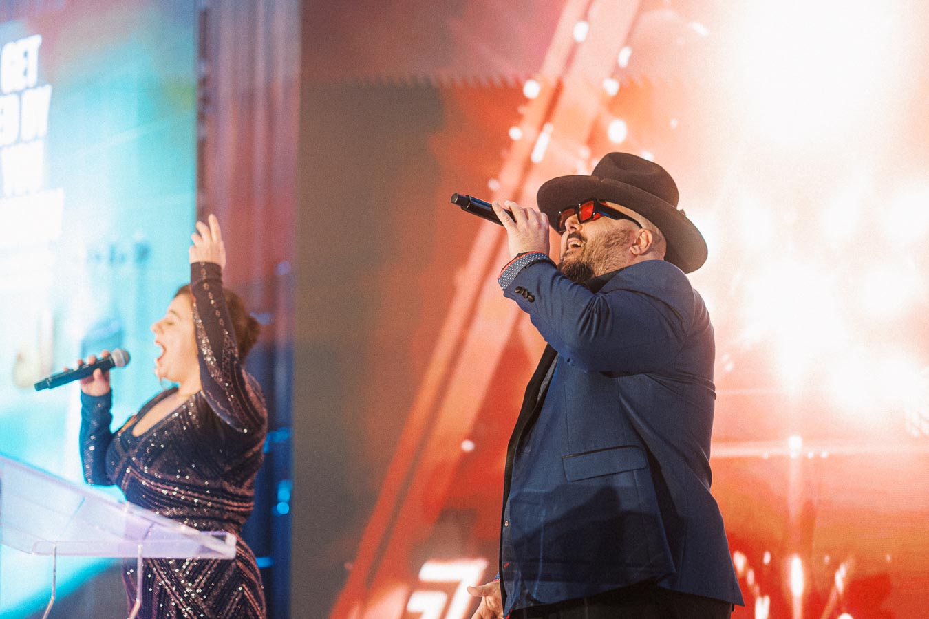 Performers on stage singing passionately at a vibrant event with colorful lights and dynamic atmosphere.