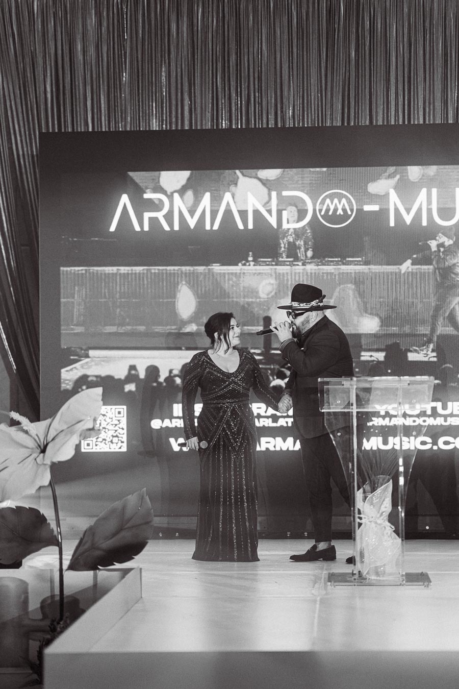 Black and white image of two performers on stage at an event, with one holding a microphone. A large screen displaying Armando Music is in the background, along with QR codes and a website URL. Decorative plants are visible in the foreground.
