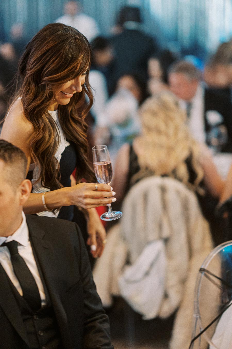 A woman holding a glass of champagne at an elegant event, with guests in formal attire in the background, creating a sophisticated and celebratory atmosphere.