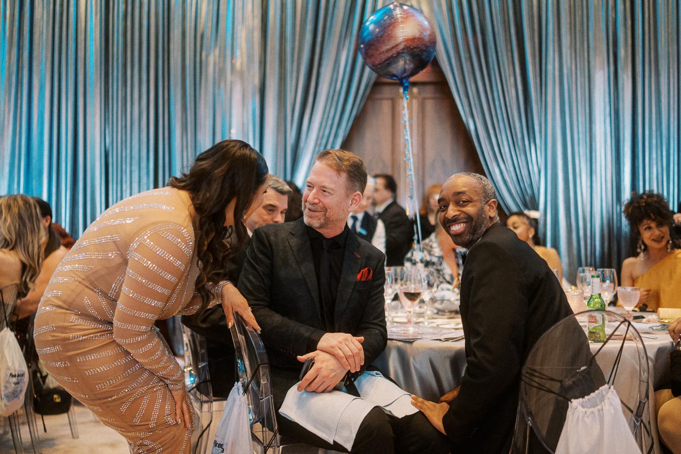 A group of well-dressed people enjoying a conversation at an elegant indoor event with a metallic blue backdrop and a decorative balloon.