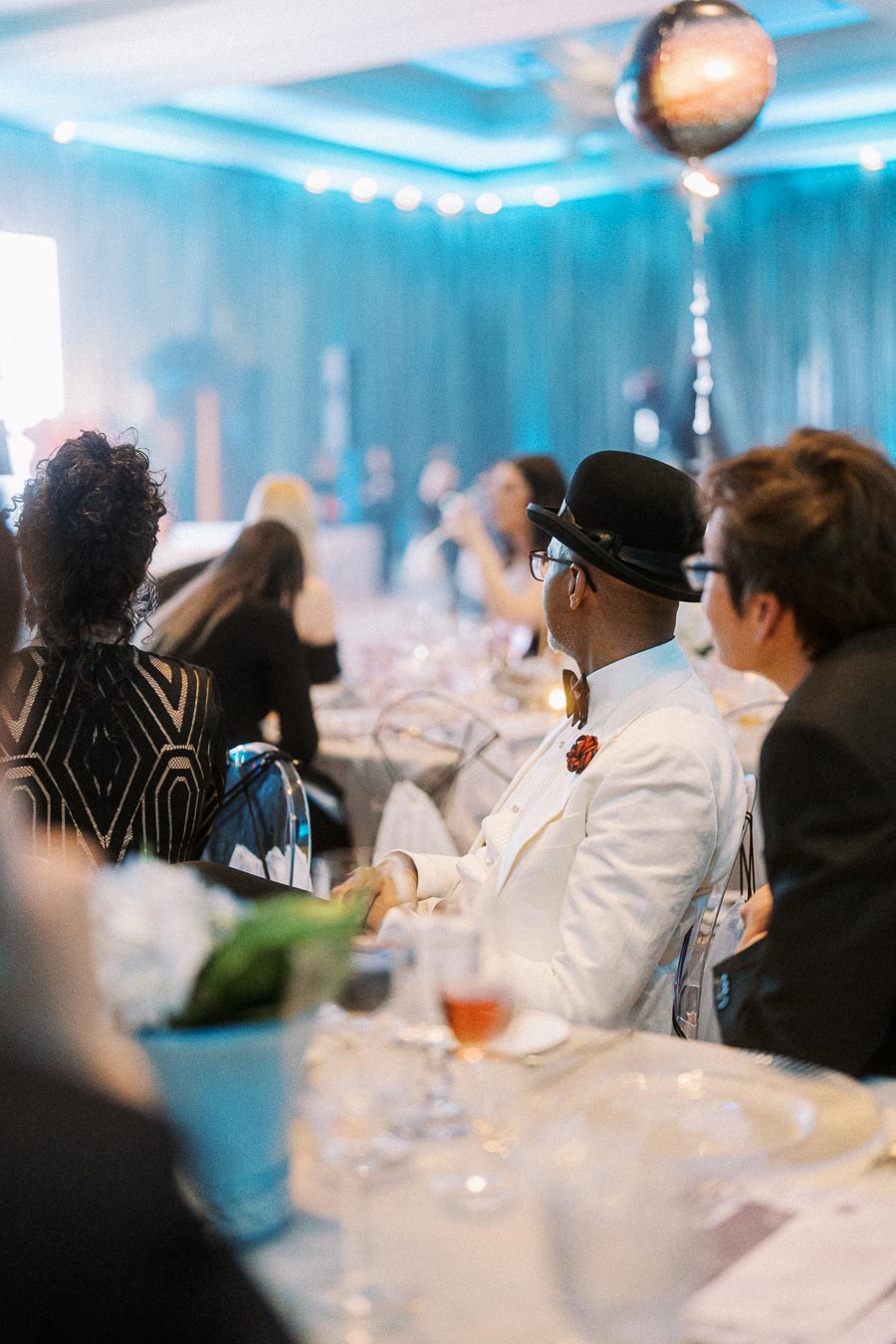 Guests seated at an elegantly set table during a formal event, with a focus on the stylish attire of attendees, under ambient lighting and event decor.