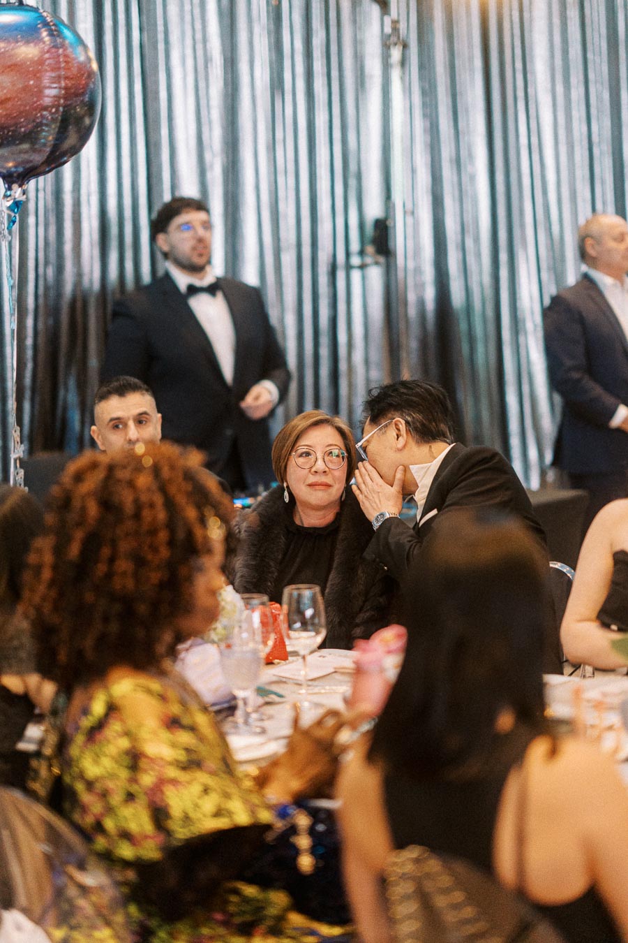 Guests at a formal event, seated at a table with elegant table settings, engage in conversation. A man in a tuxedo stands in the background, while balloons add a festive touch.