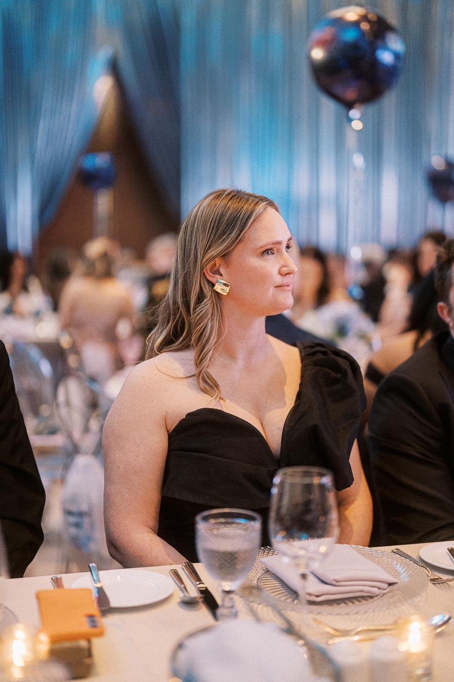 A woman in an elegant black dress attentively listens at a formal event, seated at a table with fine dining settings, surrounded by other guests in a luxurious, dimly lit venue.