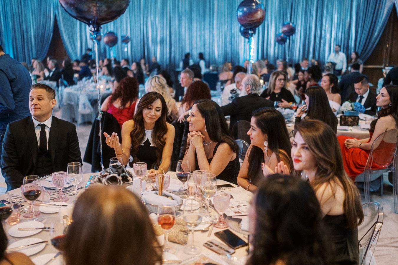 Elegant dinner event with guests in formal attire seated at a beautifully set table, featuring wine glasses and decorative balloons, against a stylish blue draped background.