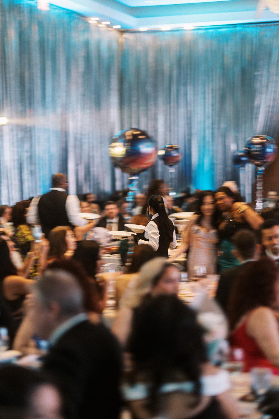A bustling event with blurred guests seated at tables, servers carrying plates, and elegant balloon decorations creating a festive atmosphere.