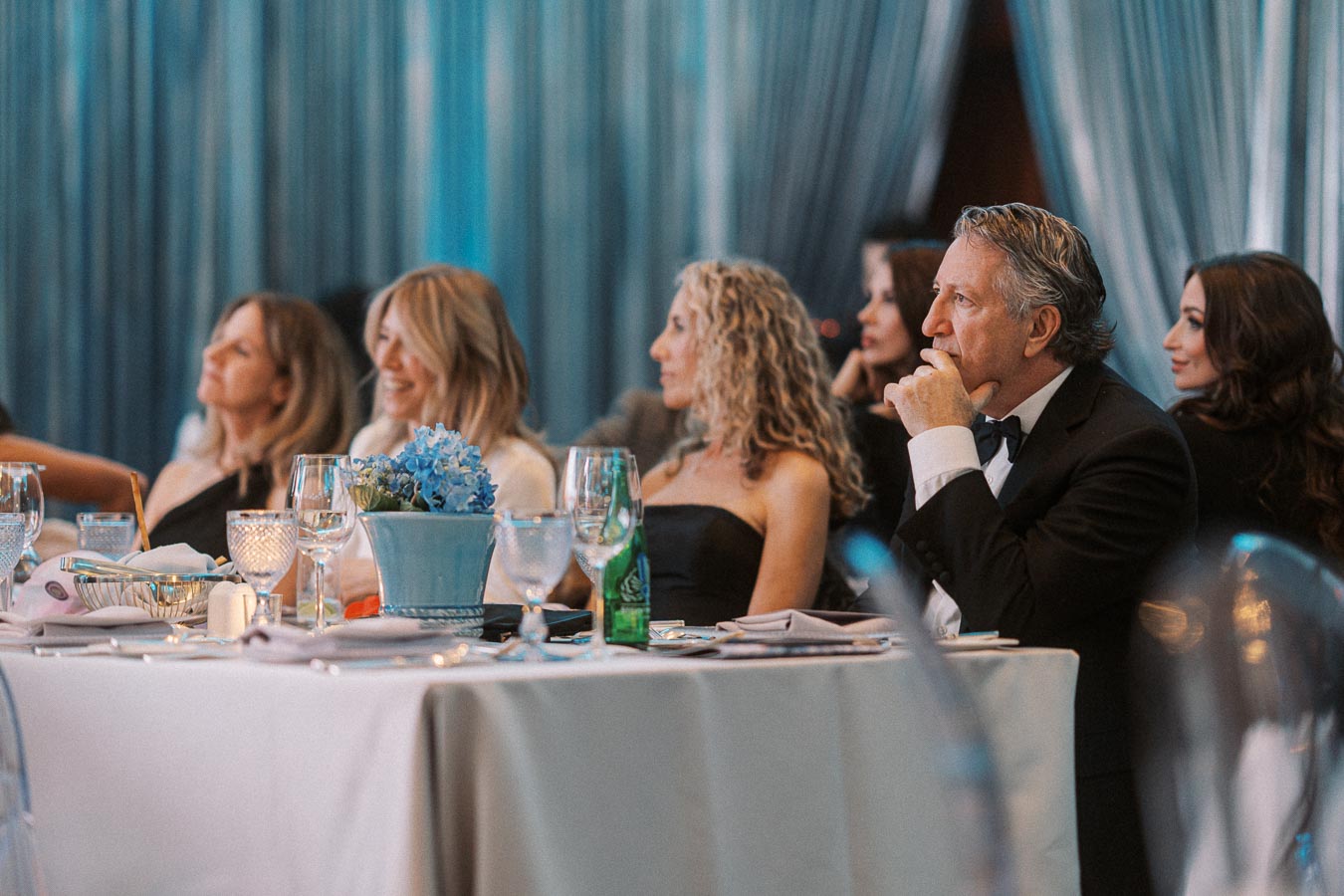 Group of elegantly dressed people seated at a banquet table, attentively listening at a formal event, with blue drapery and a centerpiece of blue flowers.