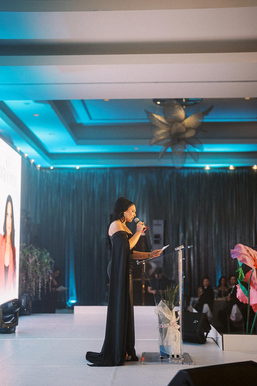A woman in an elegant black dress speaks into a microphone on stage at a sophisticated event, with a podium and a large floral decoration, under ambient blue lighting.