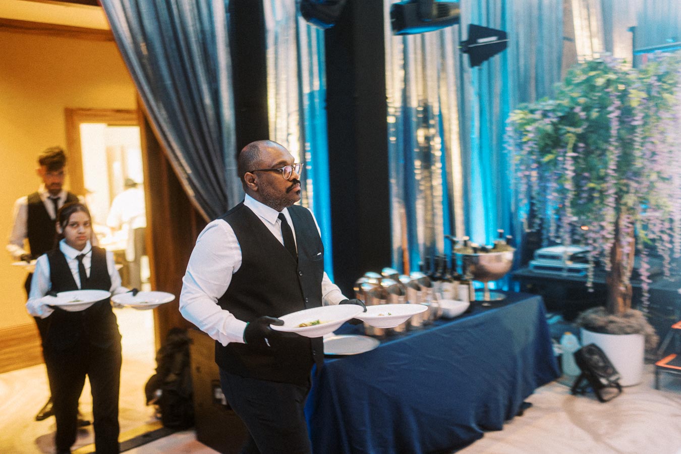 Waitstaff in formal attire carrying plates at an elegant event with blue decor and ambient lighting.