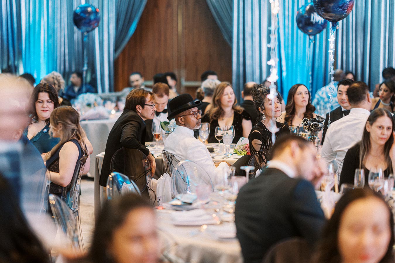 Elegant group of people seated at a formal banquet event, surrounded by decorative balloons and a beautifully set table with dinnerware and glasses, under a blue-lit ambiance.