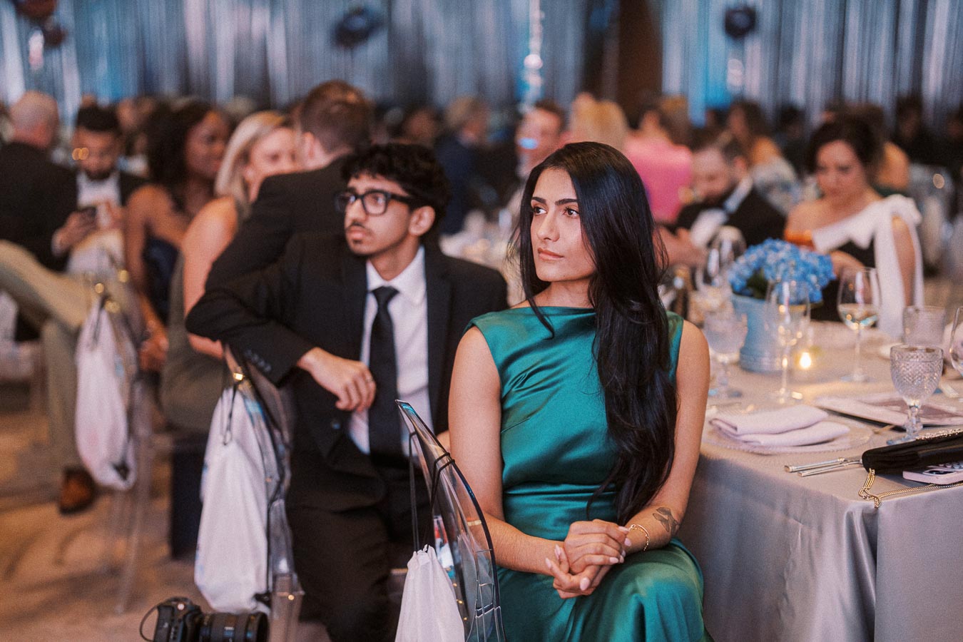 Elegant evening event with people seated at a formal table, featuring a woman in a teal dress attentively listening, surrounded by well-dressed guests in a softly lit atmosphere.