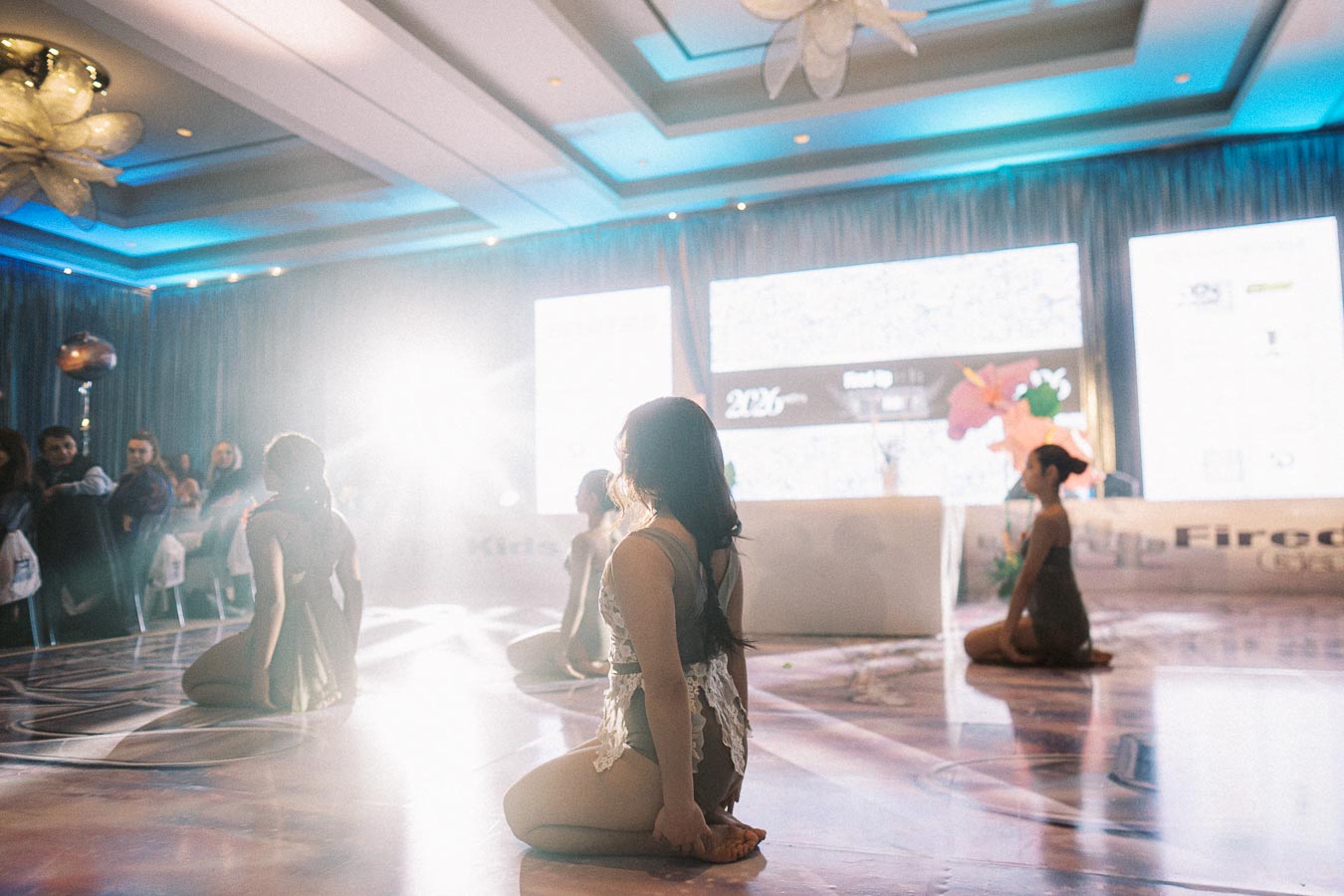 Dancers performing in a well-lit ballroom with an audience in the background, featuring elegant stage lighting and modern decor.