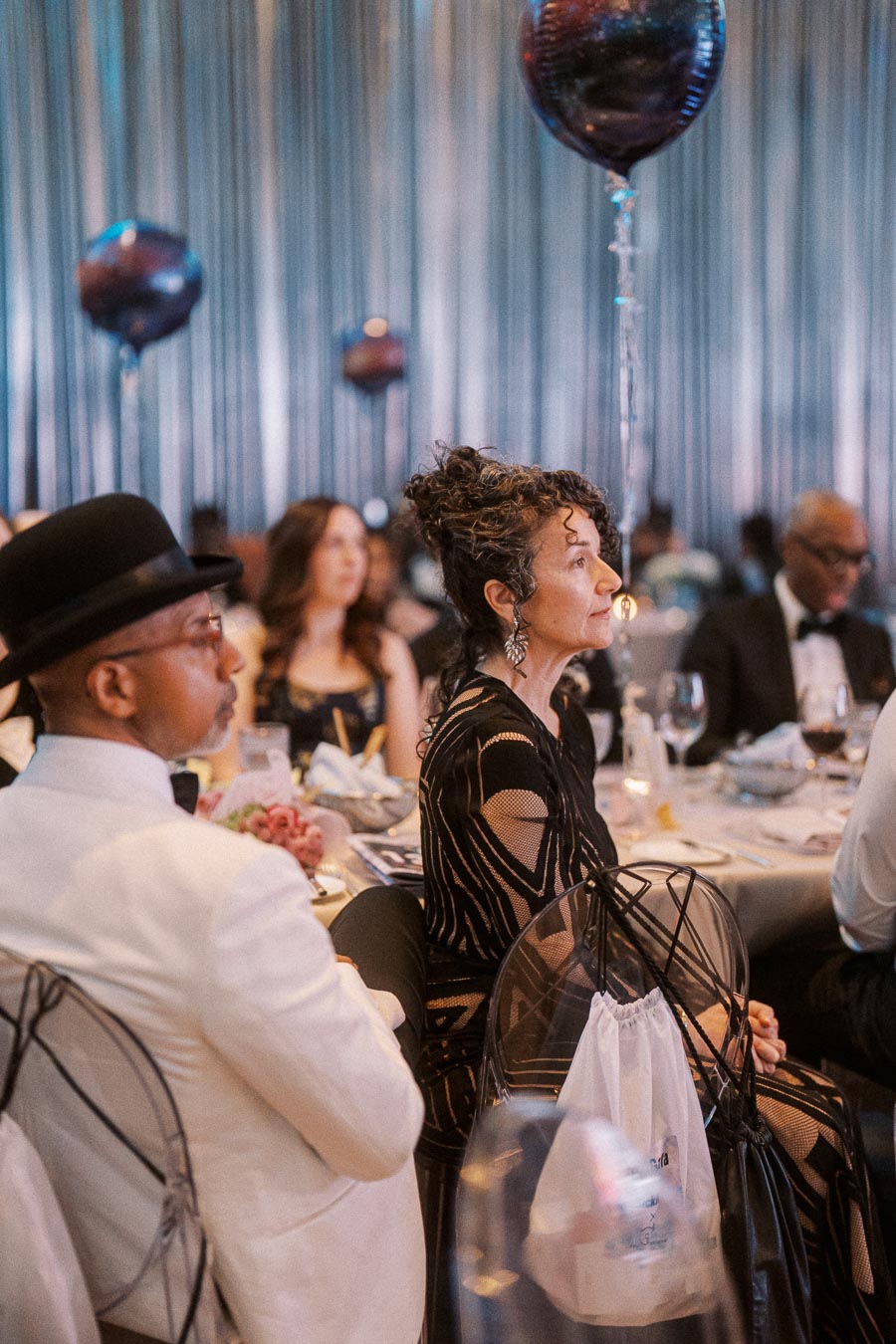Elegant guests seated at a formal dinner event, featuring sophisticated attire and festive balloons in the background, creating a refined and celebratory atmosphere.