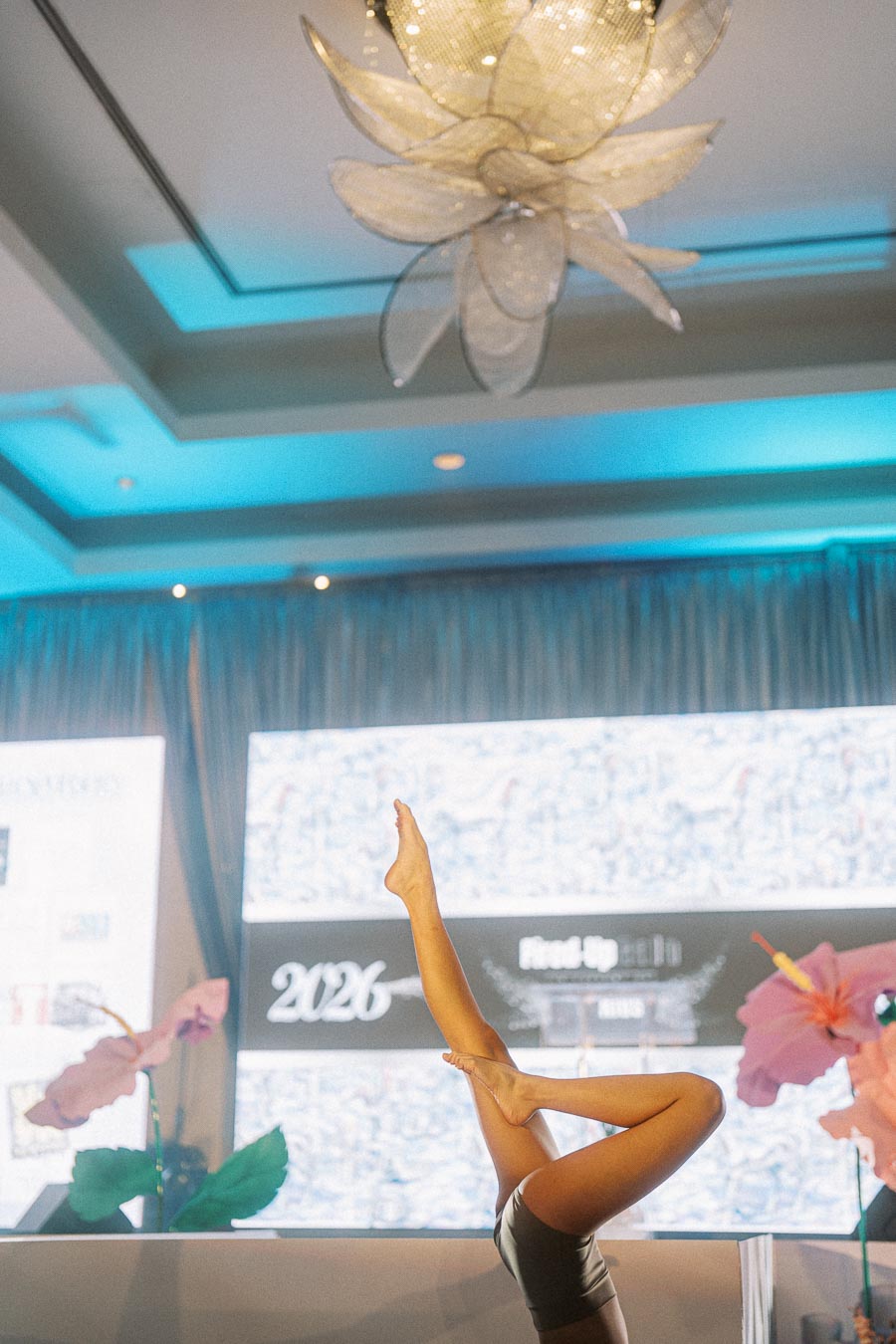 Aerial view of a modern indoor event space featuring an elegant chandelier and a performer executing a dynamic leg pose on stage, with vibrant floral decorations and multimedia displays in the background.