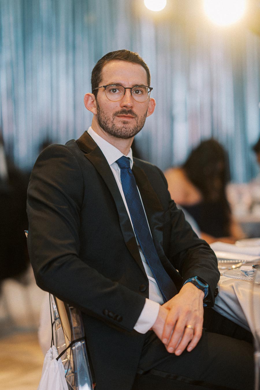 A man wearing a black suit and glasses sitting at a formal event table, with a blurred background and warm lighting.