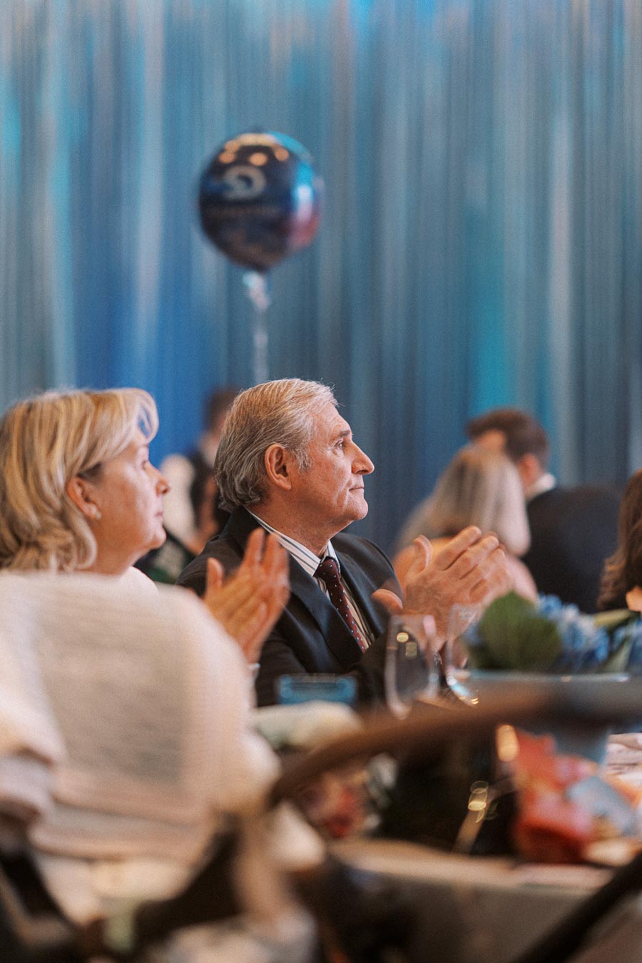 Attendees at a formal event clapping, with a man and woman in focus, and a balloon in the background, set against a blue draped backdrop.