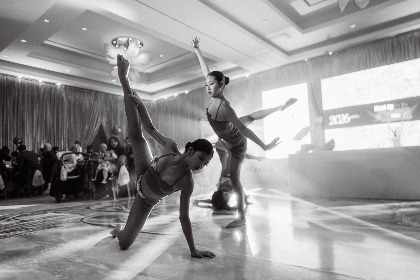 Black and white photo of three contemporary dancers performing an elegant routine at an indoor event, showcasing their flexibility and grace on a polished floor, with an audience in the background.