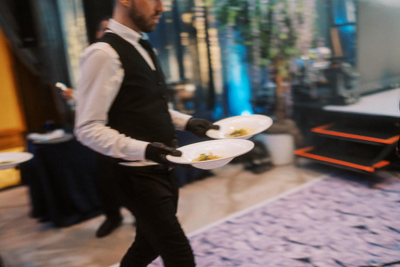 Waiter in a formal uniform carrying plates with prepared dishes at a restaurant event.