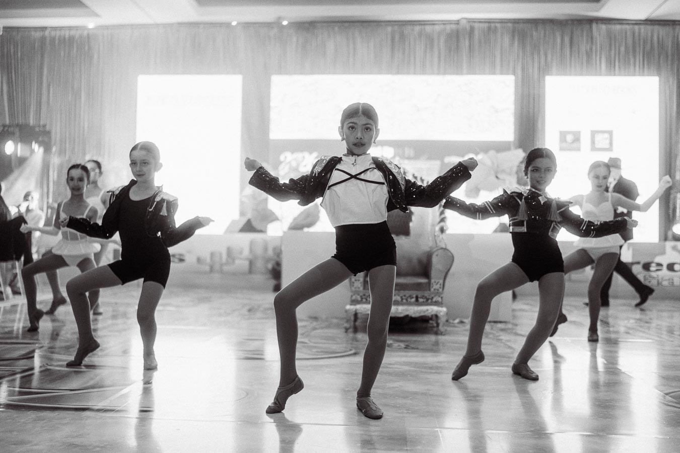 Young dancers performing a choreographed routine indoors, wearing coordinated outfits. The black and white image captures the energy and precision of the dance group as they strike dynamic poses on a polished floor.