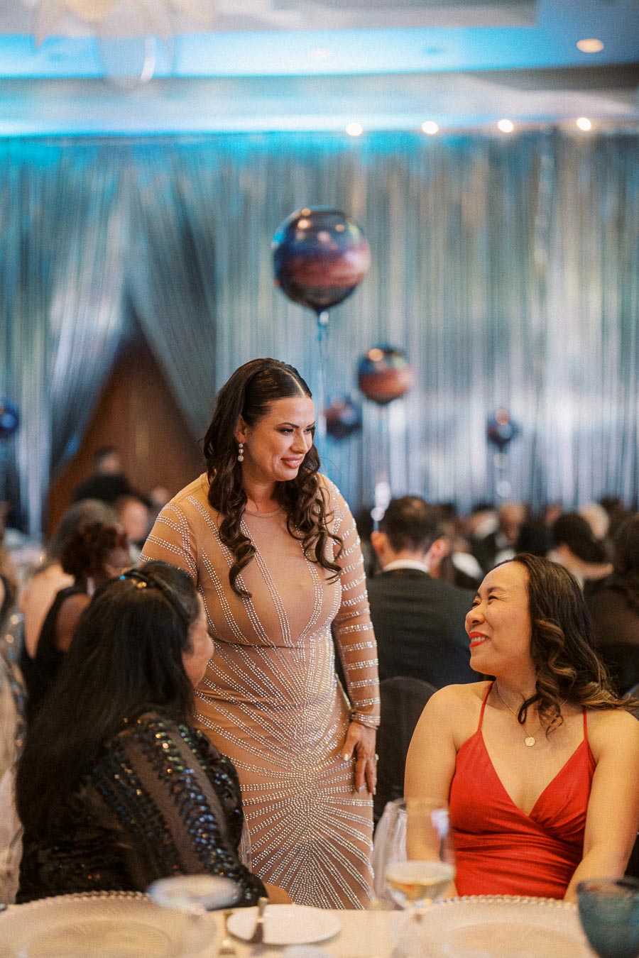 A woman in an elegant, sparkling evening gown socializes with two seated women during a formal event, with blue and silver decor in the background, creating a sophisticated atmosphere.