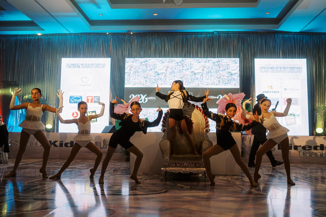 A group of young dancers in elegant costumes perform a choreographed routine on a stage with a backdrop of sponsor logos at a vibrant event.