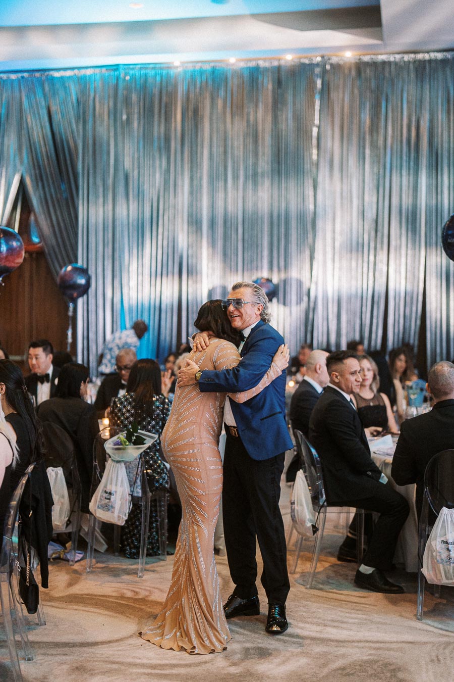 A joyful moment at a glamorous event as two elegantly dressed people embrace amidst seated guests, with a shimmering curtain backdrop and balloon decorations.