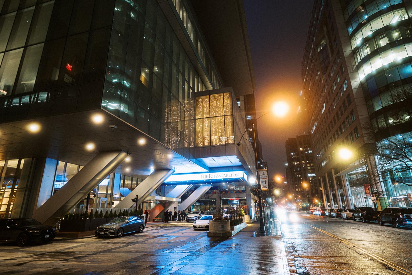 Exterior view of a modern luxury hotel at night, featuring illuminated streetlights and cars parked on a wet city street.