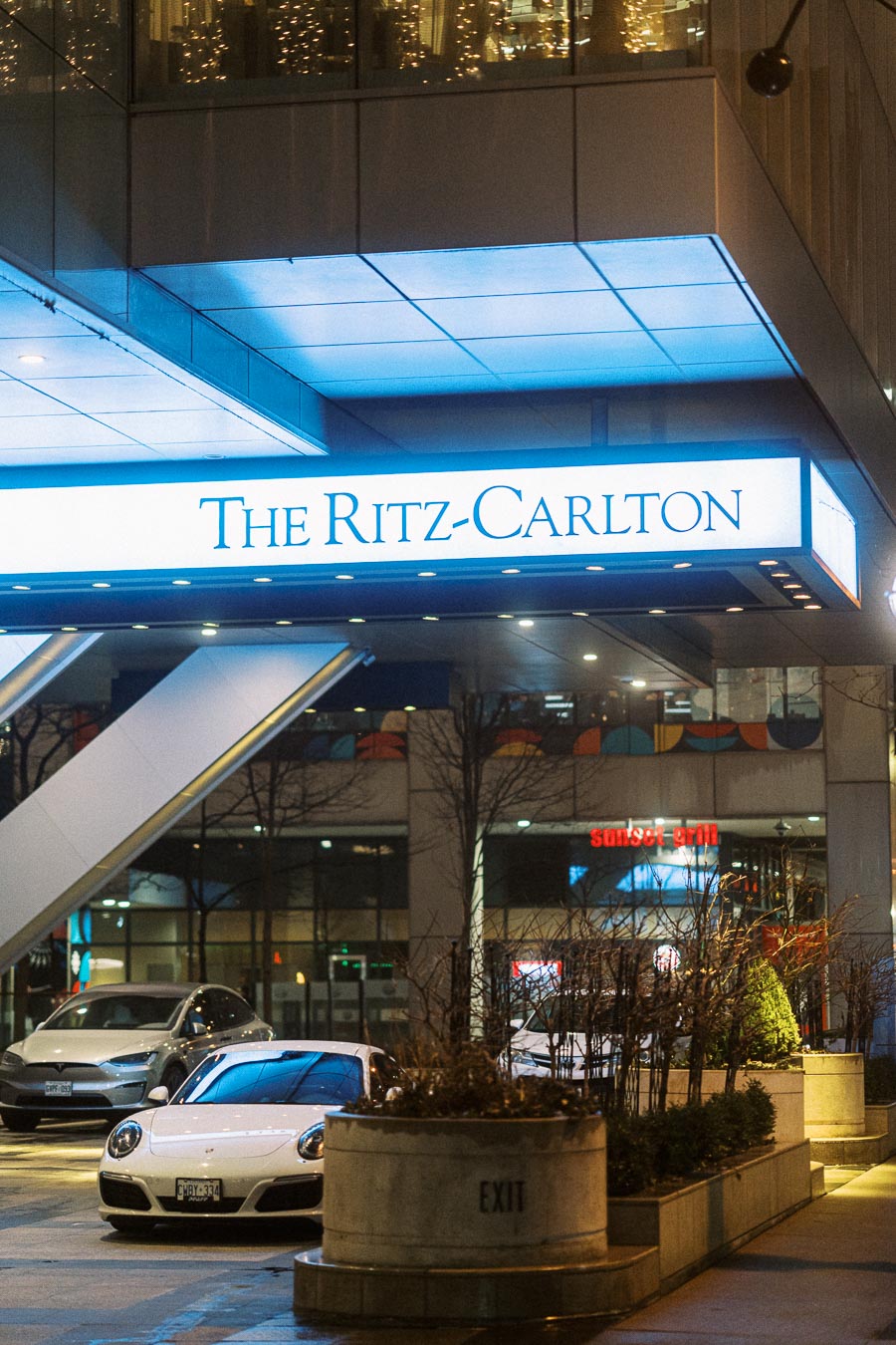 Exterior view of The Ritz-Carlton hotel entrance at night, featuring luxury cars parked in the foreground and modern architecture illuminated by city lights.