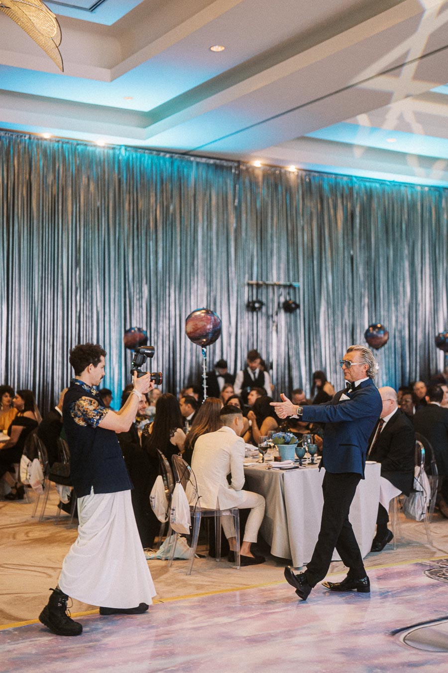 Formal event with guests seated around tables, a photographer capturing moments, and a person in a blue suit walking on a decorated floor. Elegant setting with metallic draped backdrop and creative lighting.