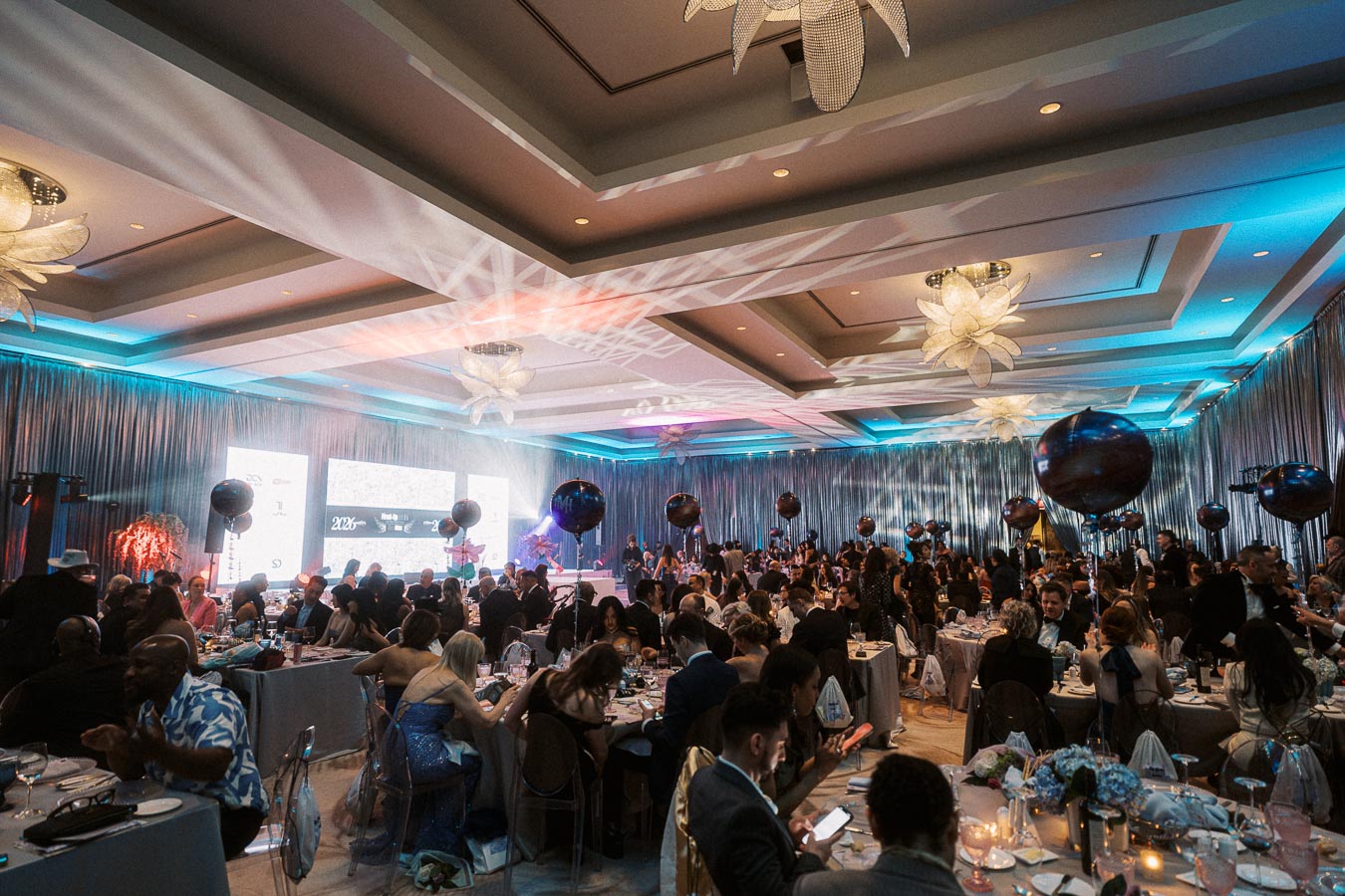 A lively and elegant gala event with guests seated at beautifully decorated tables in a spacious venue, featuring ambient blue lighting and decorative balloons throughout the room.