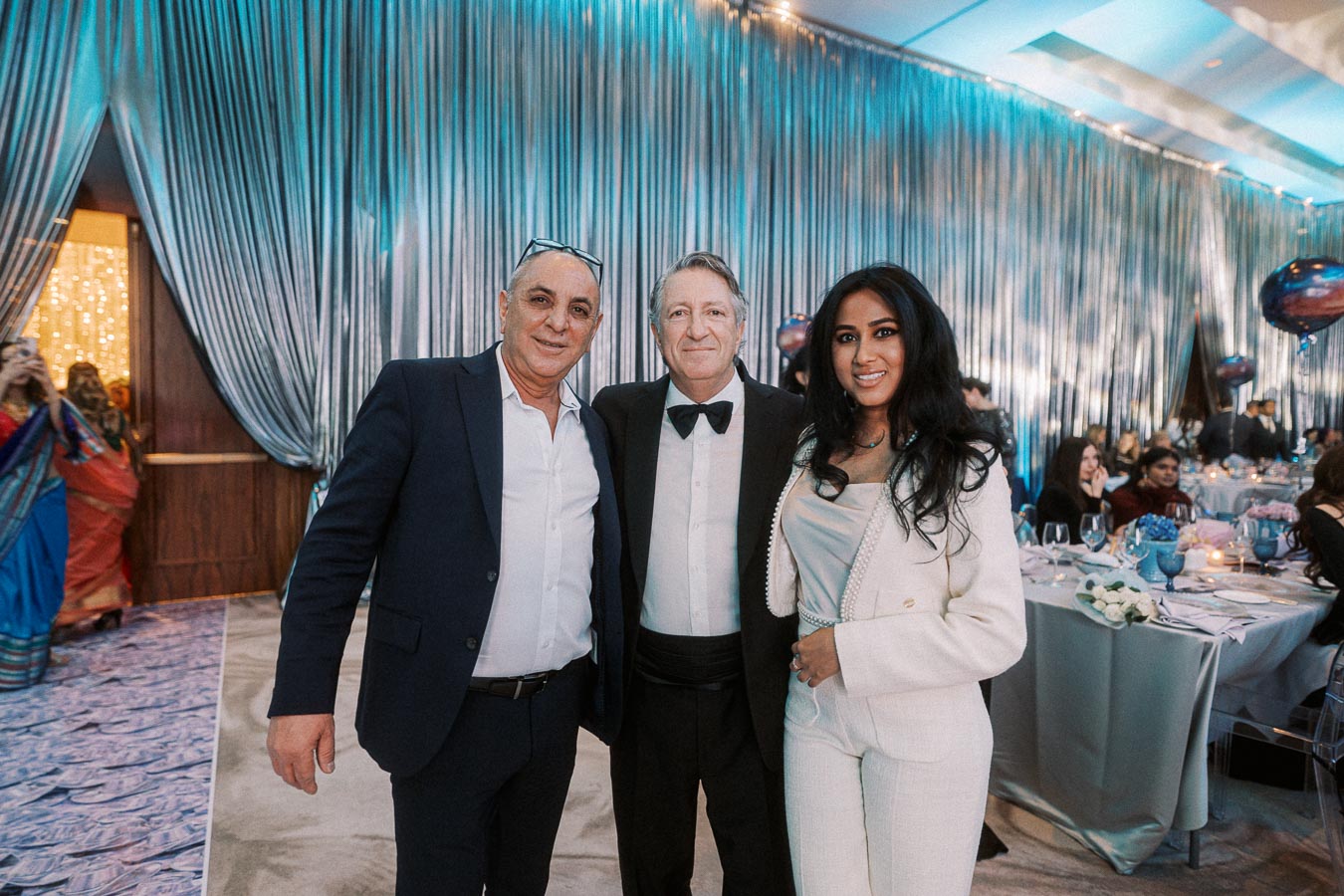 A group photo at an elegant event, featuring three people dressed in formal attire, posing together in a beautifully decorated venue with blue drapes and ambient lighting. The background shows banquet tables set for a formal dinner with guests seated.