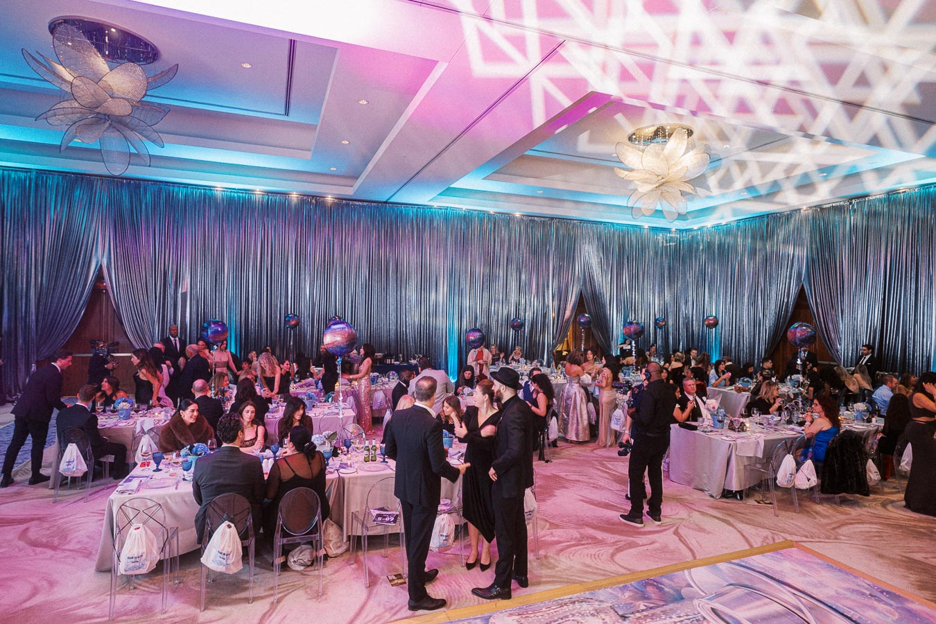 A glamorous gala event inside a luxurious ballroom decorated with shimmering curtains and colorful lighting, featuring elegantly dressed guests mingling and seated at beautifully set tables.