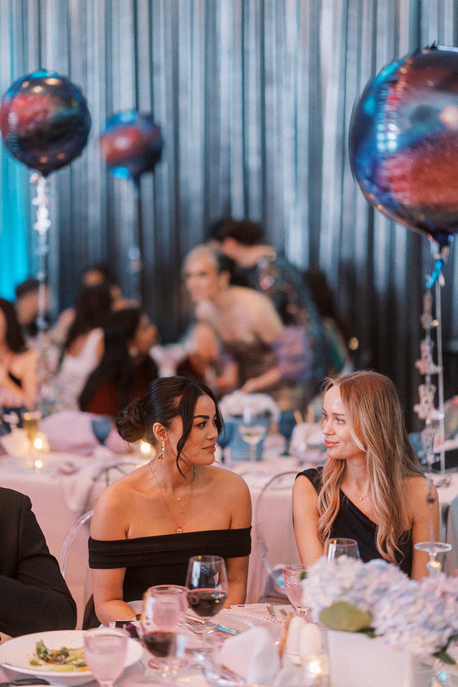 Elegant evening event with two women seated at a beautifully decorated table, surrounded by large colorful balloons and soft ambient lighting, creating a festive atmosphere.