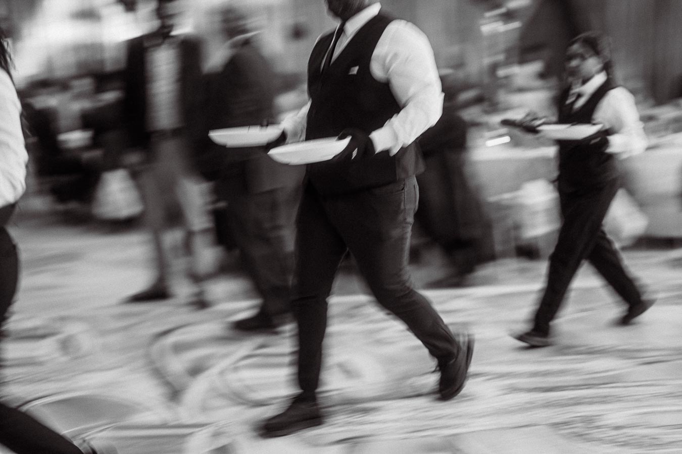 Blurred black and white photo of waitstaff in uniforms serving dishes at a busy event.