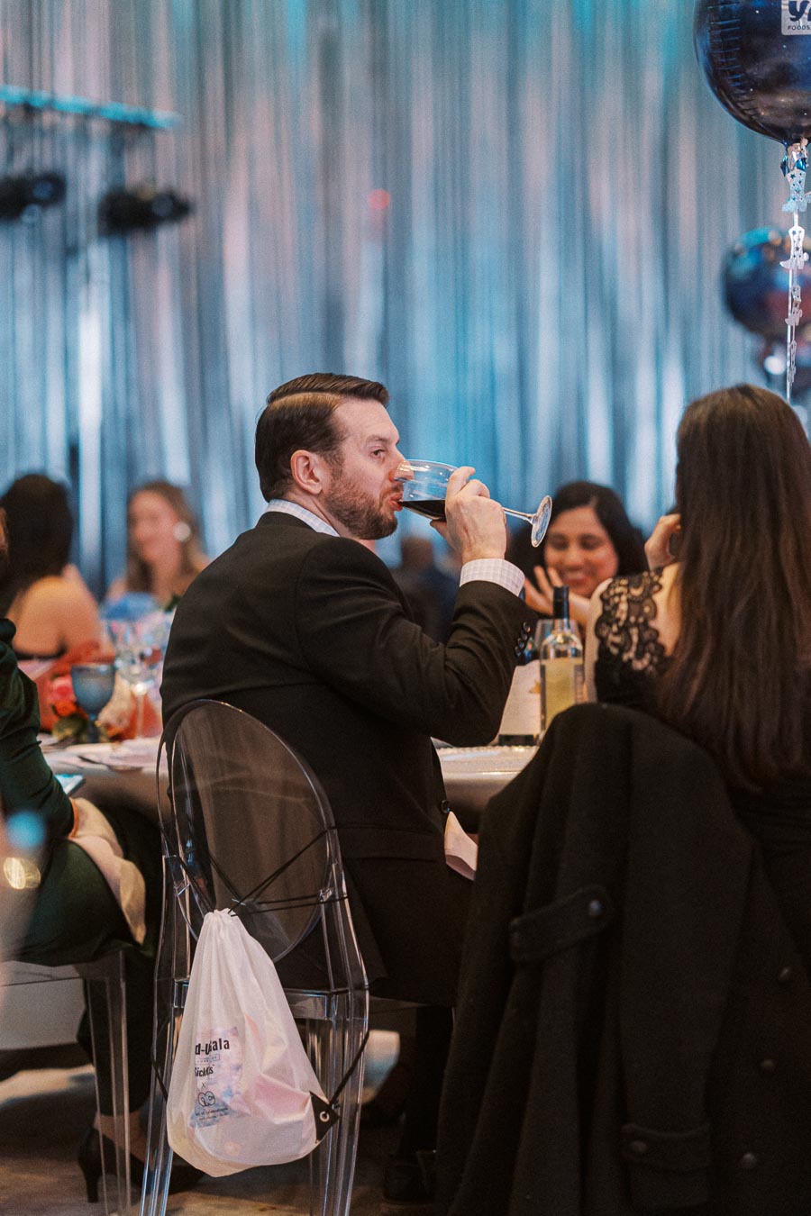 A man in a suit enjoys a glass of wine at a formal event, seated at a round table with other elegantly dressed attendees, with festive decorations and dim lighting in the background.