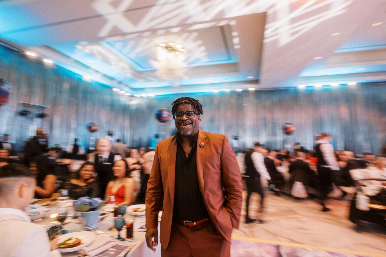 A person in a stylish brown suit smiling at a formal event with elegantly dressed attendees and a modern, well-lit banquet hall in the background.
