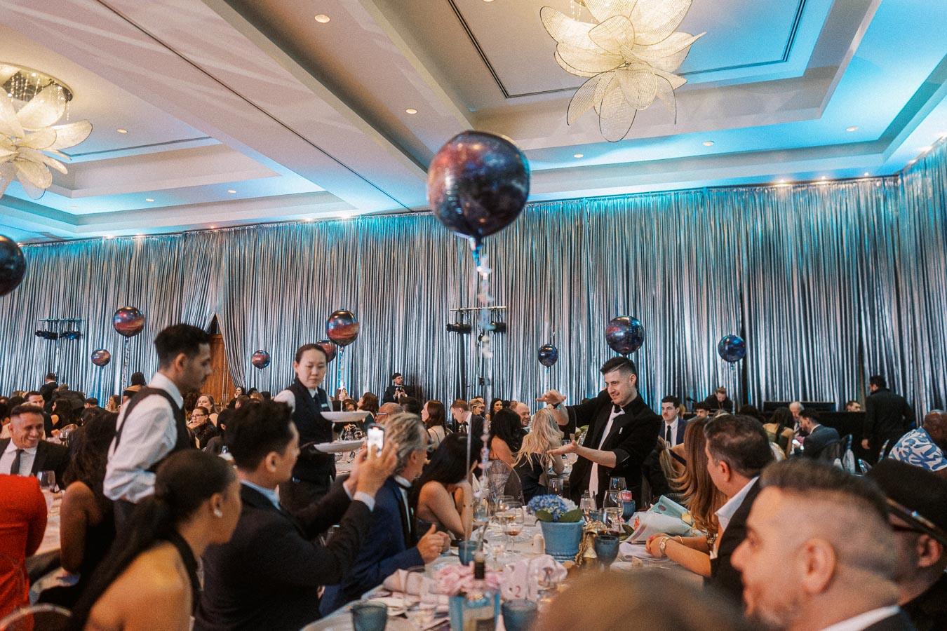 Elegant event with guests seated at decorated tables, featuring balloon centerpieces and attentive waitstaff in a luxurious venue with draped walls and modern lighting.