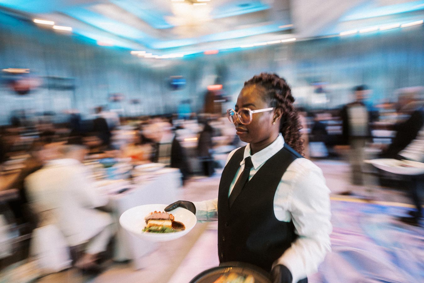 Waitstaff expertly balancing plates of gourmet food at a bustling upscale event, showcasing professional service in a lively banquet setting.