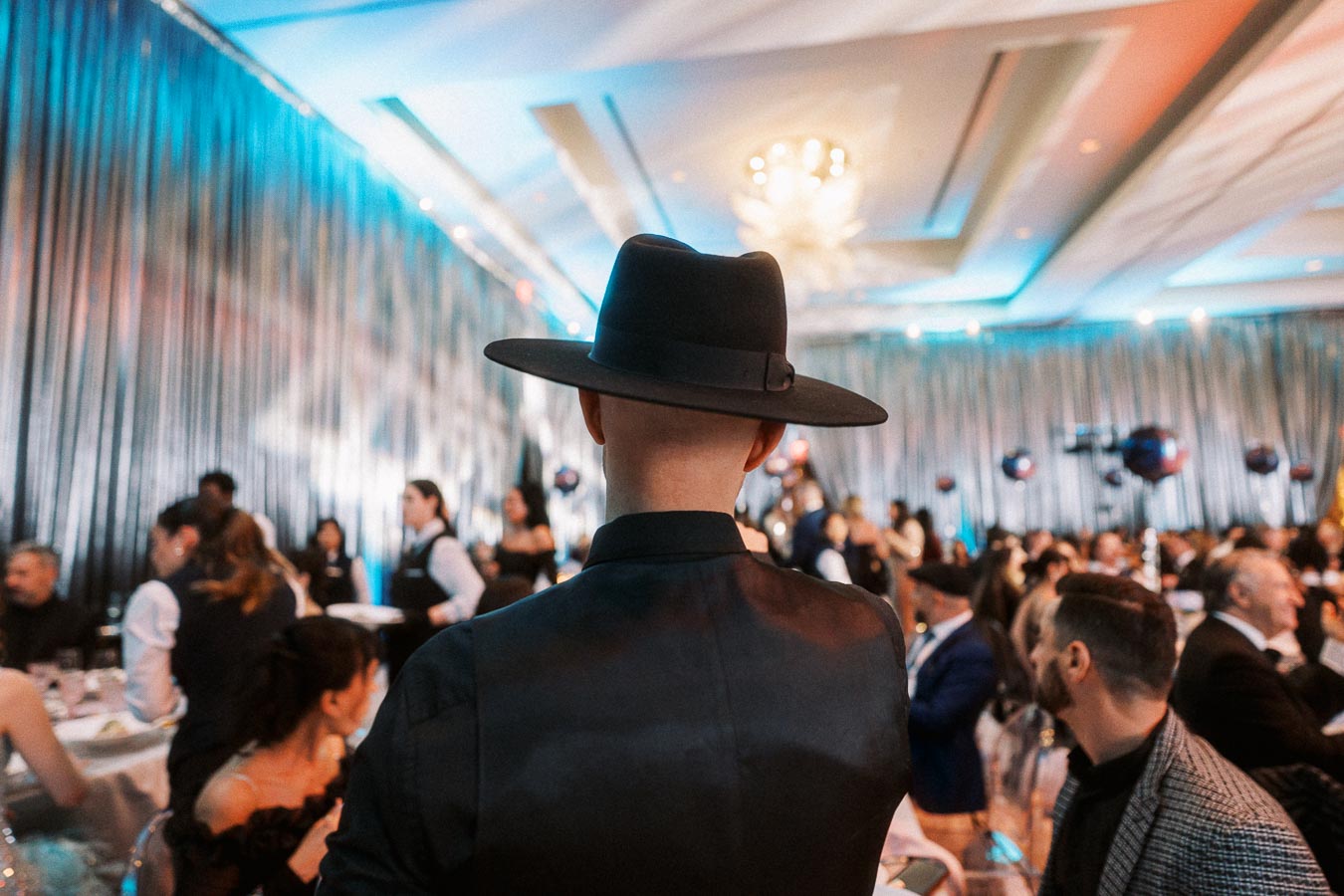 A person wearing a black wide-brimmed hat and a dark shirt stands in a dimly lit, elegant event venue, surrounded by seated guests and waitstaff, with shimmering curtains and atmospheric lighting creating a festive ambiance.